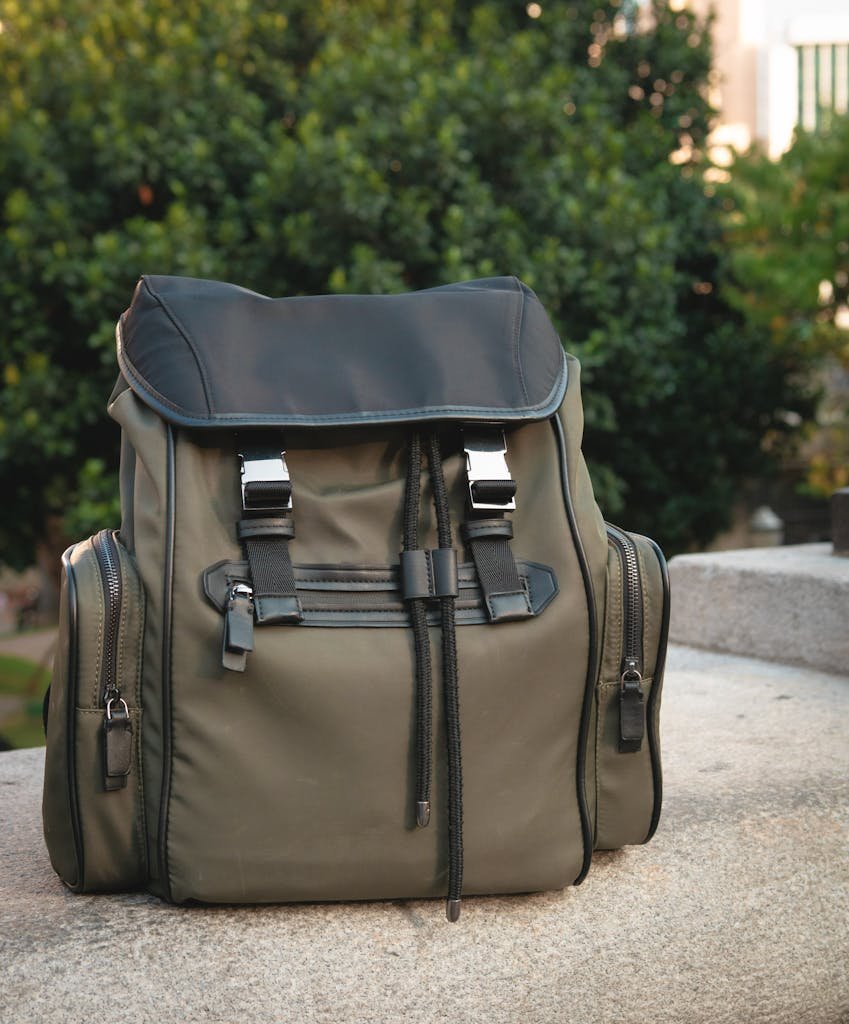 Modern olive green backpack with leather details sitting outdoors on stone. Perfect for travel or fashion look.